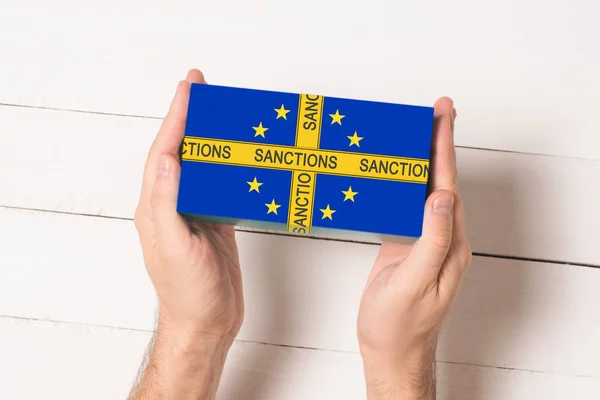 Fita amarela com a inscrição Sanções na caixa com a bandeira da União Europeia em mãos masculinas. Vista superior Mesa branca no fundo — Fotografia de Stock