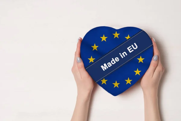 Inscrição Made in EU, a bandeira da UE. Mãos femininas segurando uma caixa em forma de coração. Fundo branco. Lugar para texto — Fotografia de Stock