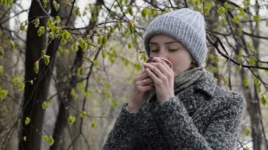 Kağıt bardaktan içen genç bir kadın. Çiçek açan yapraklar. Erken ilkbahar, karla karışık