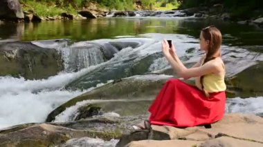 Kayanın üzerinde oturan ve selfie bir dağ nehir çağlayan arka plan üzerinde yapma Kırmızı etekli güzel uzun saçlı kız. Yaz gecesi