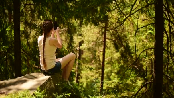 Menina bonita sentada em uma pedra e fazendo selfie e filma vídeo em um smartphone. Floresta de coníferas, dia de sol . — Vídeo de Stock