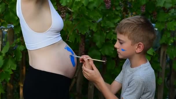 Gülen çocuk, hamile annesinin bebeğinin ayak izinin karnına bir fırça yla çizer. Hamile kavramı. Close — Stok video