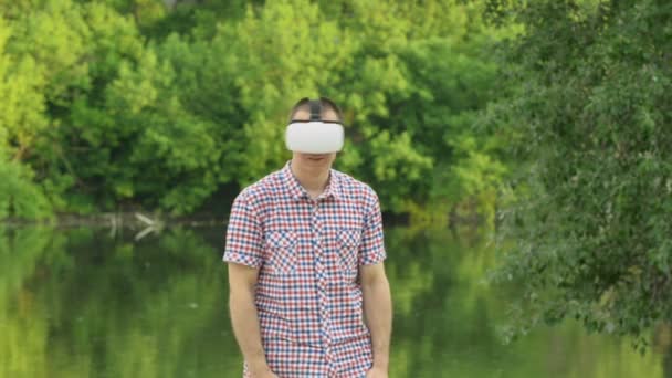 Man maakt gebruik van een virtual reality helm buitenshuis. Oever van de rivier, zomerdag in de natuur. Technologie in de buitenlucht — Stockvideo