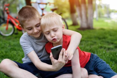 Akıllı telefon kullanan iki çocuk çimlerin üzerinde oturuyor. Arka planda bisikletler