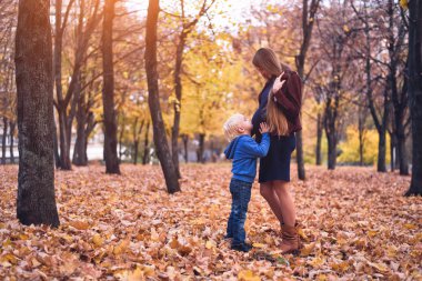 Küçük sarışın çocuk hamile annesinin karnına sarılıyor. Arka planda sonbahar Parkı