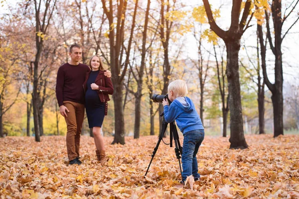 Liten blond pojke med en stor SLR-kamera på ett stativ. Fotografier ett gift par, graviditet. Familjefotosession — Stockfoto