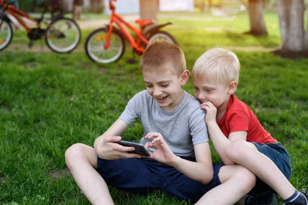 Çimlerin üzerinde otururken iki çocuk akıllı telefonda oyun oynuyor. Arka planda bisikletler