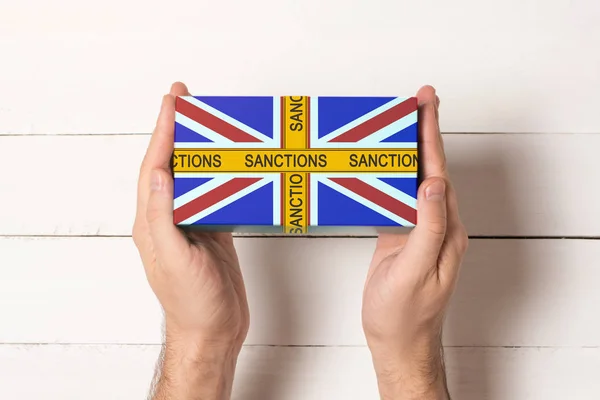 Žlutá páska s nápisem sankce na krabici s vlajkou Spojeného království v rukou mužů. Pohled shora bílá tabulka na pozadí — Stock fotografie