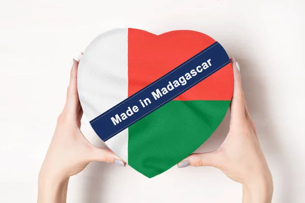 Felirat Made Madagaszkáron, a zászló Madagaszkár. Egy szív alakú dobozt tartó női kéz. Fehér háttér. — Stock Fotó