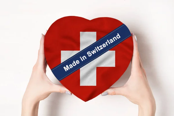 Inscripción Hecho en Suiza la bandera de Suiza. Manos femeninas sosteniendo una caja en forma de corazón. Fondo blanco . —  Fotos de Stock