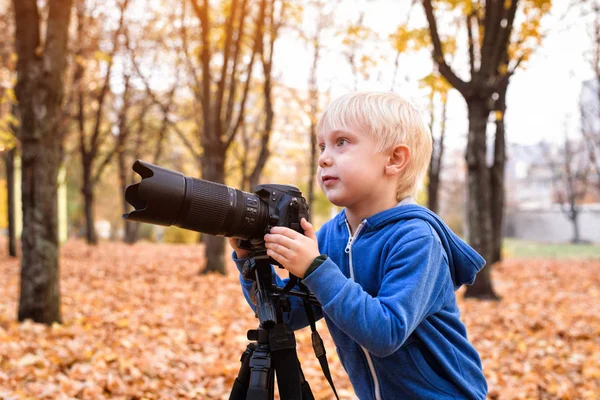 小さなブロンドの男の子は、一眼レフカメラで写真を撮ります。秋の公園 — ストック写真