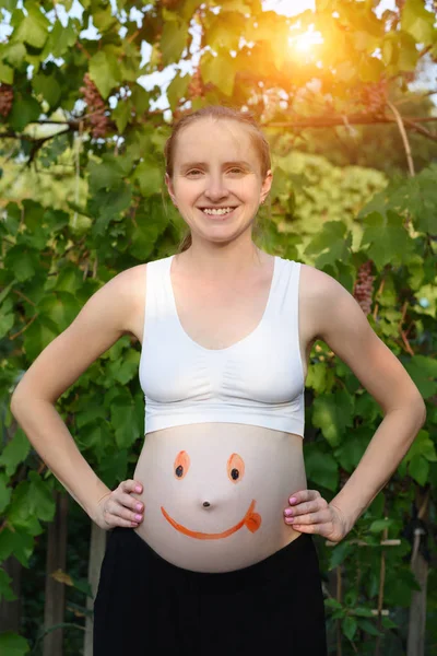 Jovem grávida sorridente com um rosto sorridente pintado na barriga. Fundo verde — Fotografia de Stock