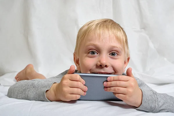 小さなブロンドの男の子は、彼の手にスマートフォンを持ってベッドに横たわっています。ガジェットレジャー — ストック写真