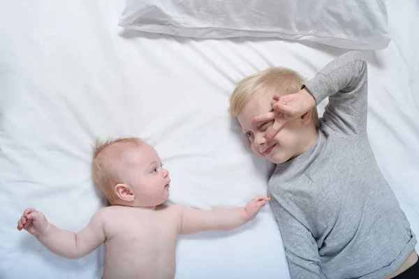 Bebé y hermano mayor sonriente están acostados en la cama. Juegan, son graciosos e interactúan. Vista superior —  Fotos de Stock