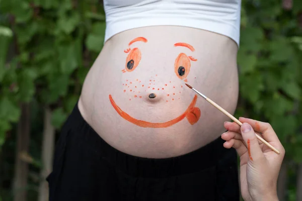 Ręka z pędzlem farby na brzuchu kobiety w ciąży uśmiechnięta twarz. Zielone tło — Zdjęcie stockowe