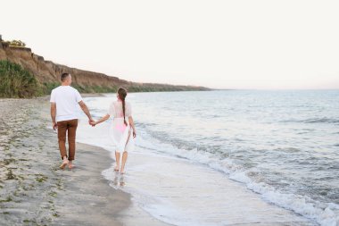 Genç çift deniz kıyısında el ele yürür. Geri görünüm