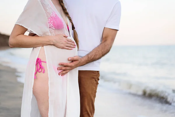 Las manos masculinas y femeninas abrazan a una barriga embarazada. Una joven pareja casada en la costa del mar. De cerca. —  Fotos de Stock