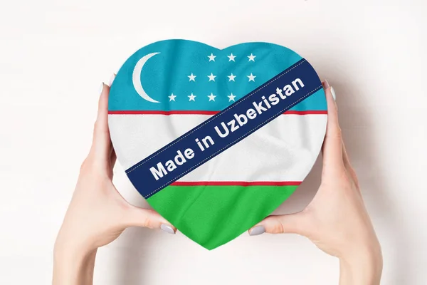 Inscription Fabriqué en Ouzbékistan le drapeau de l'Ouzbékistan. Mains féminines tenant une boîte en forme de cœur. Fond blanc . — Photo
