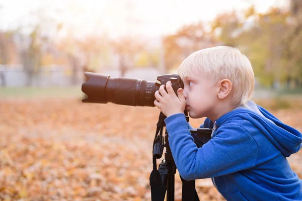 Portret małego chłopca blond z dużym aparatem DSLR na statywie. Sesja zdjęciowa w jesiennym parku — Zdjęcie stockowe