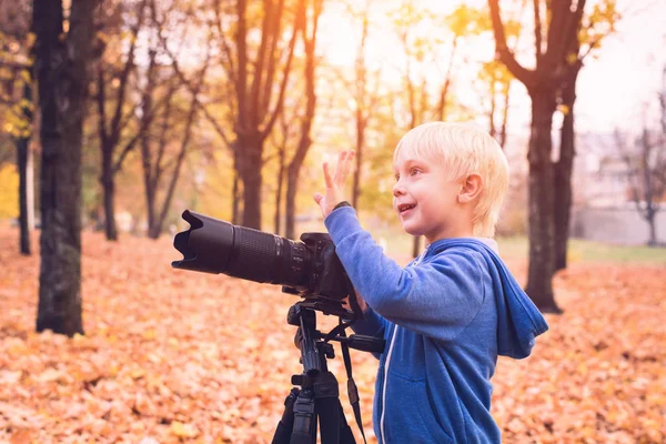 Malý blonďák střílí s velkou SLR kamerou na trojnožce. Fotografická relace v podzimním parku — Stock fotografie