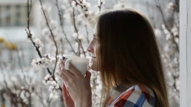 Bir balkon cam kenarında duran ve çiçek açan kayısı bir arka plan üzerinde çay içme bir ekose kız