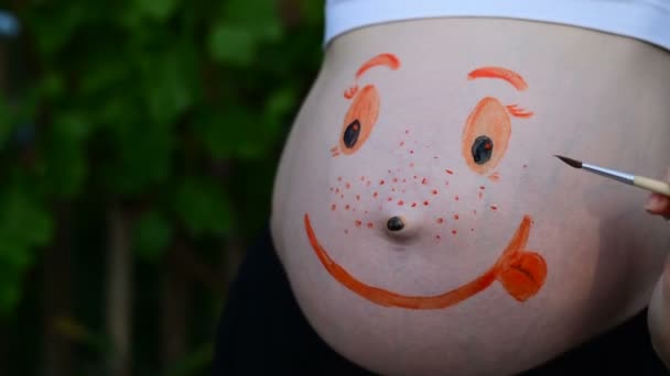 Рука с кисточкой рисует на беременном животе смешное лицо. Беременная концепция. Крупный план — стоковое видео