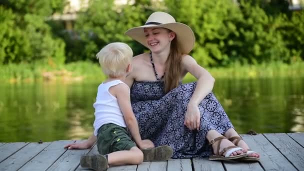 Mamá y su hijo rubio se abrazan y charlan en el muelle junto al río. Día de verano — Vídeos de Stock