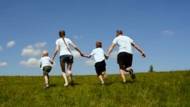 Mutlu aile anne, baba ve iki küçük oğlu mavi gökyüzü ve bulutların bir arka plan üzerinde el ele tutuşarak yeşil bir çayırda koşmak