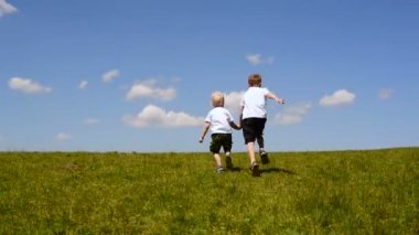 Yeşil bir çayırda koşan iki küçük çocuk mavi gökyüzü ve bulutların arka planında el ele tutuşuyor.