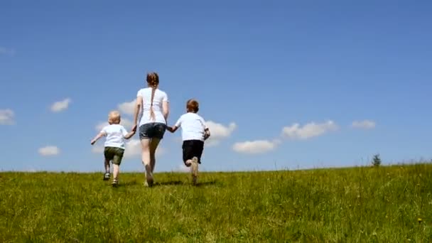 Madre y dos hijos pequeños corren a través de un prado verde tomados de las manos sobre un fondo de cielo azul y nubes — Vídeo de stock