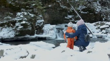 Anne ve oğlu kucaklama şelale, hill ve çam ormanlar karla kaplı bir arka plan üzerinde oturuyor. Kış bulutlu gün