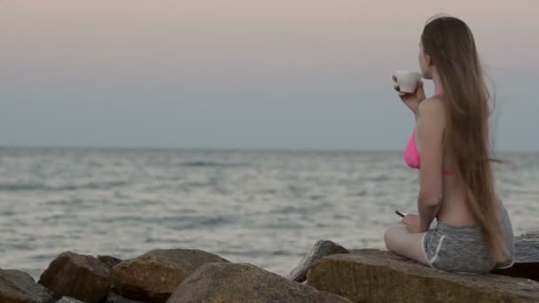Piękna dziewczyna siedzi na plaży i picie kawy z filiżanką. Czas zachodu słońca — Wideo stockowe
