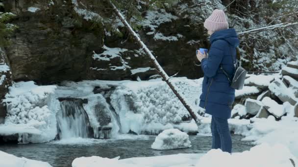 Chica de abrigo azul de pie con una taza sobre un fondo de cascada helada y rocas en un bosque de coníferas cubierto de nieve. Día nublado de invierno . — Vídeos de Stock
