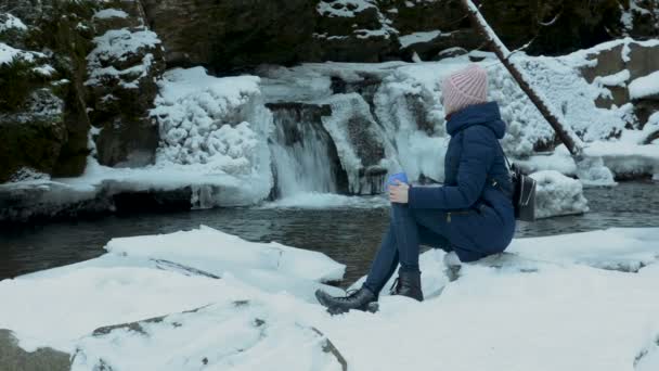 Девушка в синем пальто сидит с кружкой на фоне ледяного водопада и скал в заснеженном хвойном лесу. Зимний облачный день . — стоковое видео