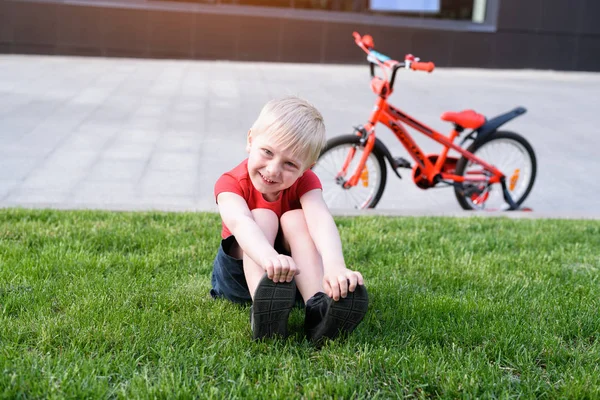 Mosolygós szőke fiú nyugszik a gyepen. Bike a háttérben — Stock Fotó