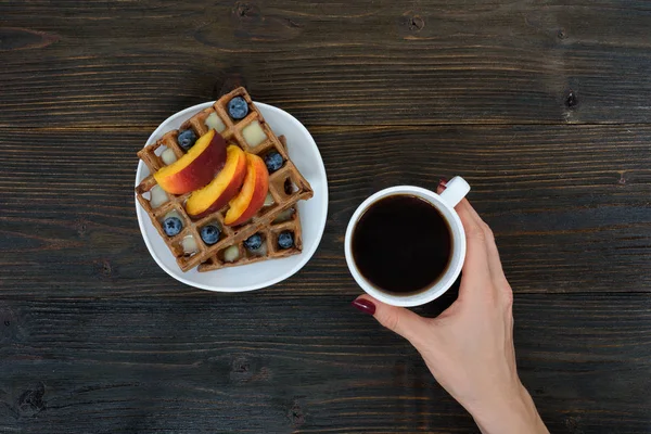 Belgische Waffeln mit Früchten und Beeren. weibliche Hand mit Tasse Kaffee. Holzuntergrund. Ansicht von oben — Stockfoto