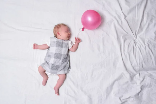 Dormir niña recién nacida en un vestido. Sábana blanca, globo rosa en la mano. Vista superior. Lugares para el texto —  Fotos de Stock