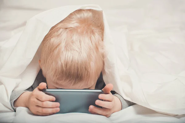 Little blond boy buried his nose in his smartphone. Lying in bed and hiding under the covers. Gadget Leisure — Stock Photo, Image