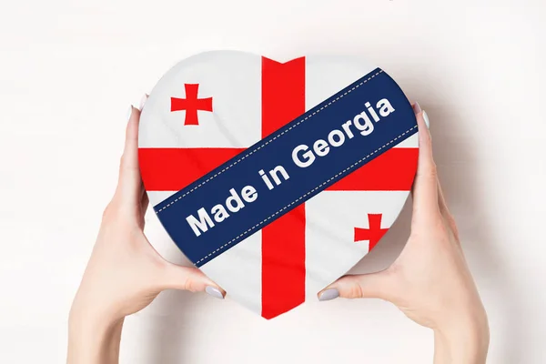Επιγραφή Made in γεωργία, η σημαία της γεωργίας. Γυναικεία χέρια που κρατάνε ένα κουτί σε σχήμα καρδιάς. Λευκό φόντο. — Φωτογραφία Αρχείου