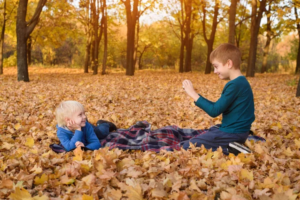 Two little brothers take pictures of each other, lying in yellow autumn leaves. Fall day — Stock Photo, Image