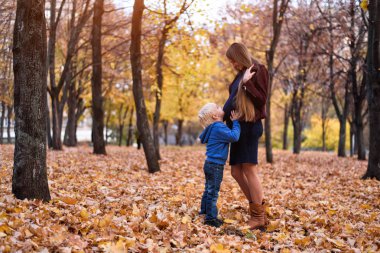 Küçük sarışın çocuk hamile annesinin karnına sarılıyor. Arka planda sonbahar Parkı