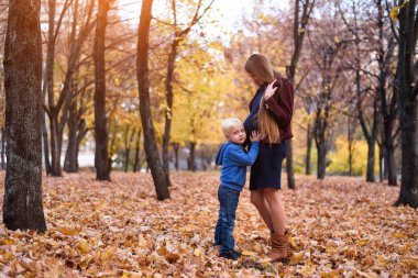 Küçük sarışın çocuk hamile annesinin karnını dinliyor. Arka planda Sonbahar parkı