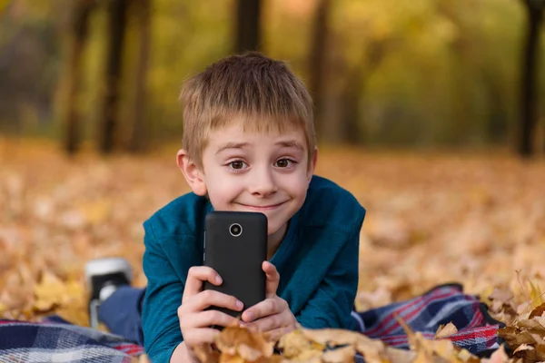 Χαριτωμένο μαθητής με smartphone βρίσκεται σε ένα καρό, κίτρινα φύλλα του φθινοπώρου. Φθινοπωρινή μέρα — Φωτογραφία Αρχείου