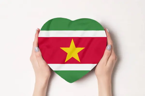 Bandeira do Suriname em uma caixa em forma de coração em mãos femininas. Fundo branco — Fotografia de Stock