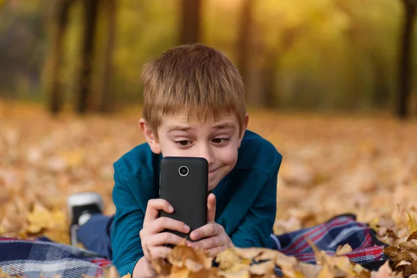 Χαριτωμένο μαθητής με smartphone παίρνει φωτογραφίες. Ξαπλωμένοι σε καρό, κίτρινα φθινοπωρινά φύλλα. Φθινοπωρινή μέρα — Φωτογραφία Αρχείου