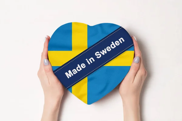 Inscripción Hecho en Suecia la bandera de Suecia. Manos femeninas sosteniendo una caja en forma de corazón. Fondo blanco . —  Fotos de Stock