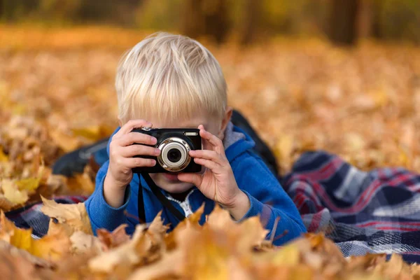 Malý blonďák s foťákem dělá fotky. Leží na plni, žluté podzimní listí. Den pádu — Stock fotografie