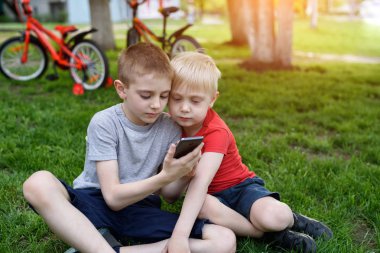Ellerinde akıllı telefon olan iki çocuk çimlerin üzerinde oturuyor. Arka planda bisikletler