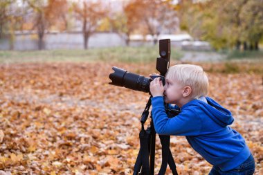 Küçük sarışın çocuk bir tripod üzerinde büyük bir SLR kamera ile vuruyor. Sonbahar parkında fotoğraf oturumu