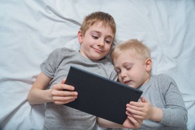 İki çocuk yatakta yatıyor ve tablette bir şey izliyor. Gadget Eğlence